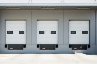 A row of three identical, closed white loading bay doors on the gray exterior of a modern warehouse. Each door has two small, horizontal windows and sits above a black dock leveler on a clean concrete lot.